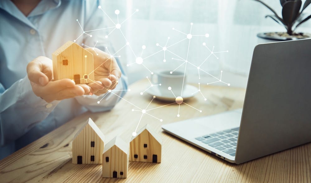 Woman holding wooden model home with other homes on desk beside laptop